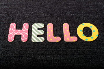 Card with Hello word made from mixed vivid colored wooden letters on a textured dark black textile material that can be used as a message.