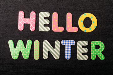 Card with Hello Winter words made from mixed vivid colored wooden letters on a textured dark black textile material that can be used as a message.