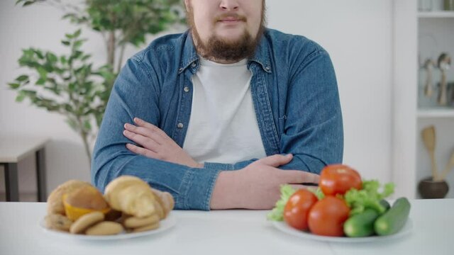 Upset plus size male looking at fresh vegetables and dessert on the table, diet