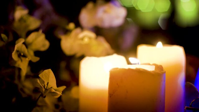 zoomed in shot of large thick wax candles with flames lit and colorful flowers all round and led lights flickering showing the table decorations at a valentine , wedding , engagement dinner at night