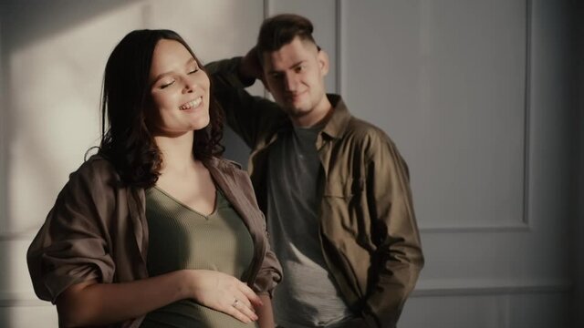 Cheerful young pregnant woman in loose clothes stands near happy gentle husband against decorated wall in room at home slow motion