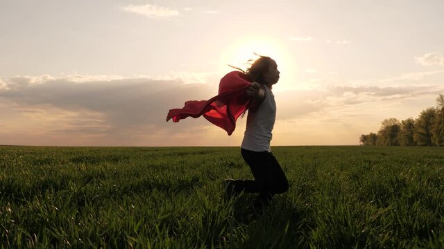 happy superhero girl, runs on green field in red cloak, cloak flutters in wind. child plays and dreams. Slow motion. teenager dreams of becoming a superhero. young girl in red cloak, dream expression.