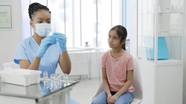 Crop view of nurse in protective medical mask and safety gloves prepering syringe with vaccine for young girl. Kid waiting for vaccination while sitting on chair in hospital.