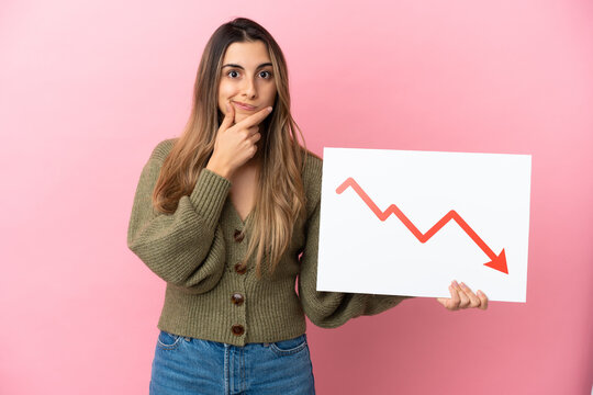 Young caucasian woman isolated on pink background holding a sign with a decreasing statistics arrow symbol and thinking