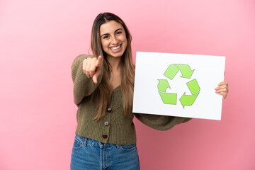 Young caucasian woman isolated on pink background holding a placard with recycle icon and pointing to the front