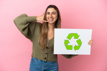 Young caucasian woman isolated on pink background holding a placard with recycle icon and doing phone gesture