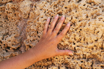 A tanned female left hand with a forearm with a gold ring on the middle finger against the background of an old weathered sand-colored stone.