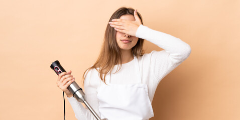 Woman using hand blender over isolated background covering eyes by hands. Do not want to see something