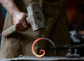 Closeup blacksmith is processing a hot metal object of a spiral shape at anvil in a workshop