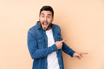 Caucasian handsome man isolated on beige background surprised and pointing side