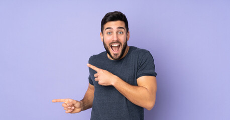 Caucasian handsome man surprised and pointing side over isolated purple background