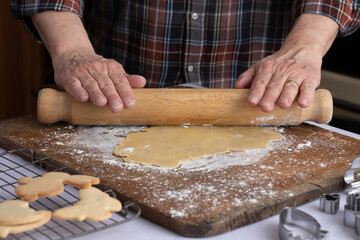 Selective focus angled semi-close-up of the hands and forearms of a person rolling dough on a wooden board with flour dusting,  some undecorated cookies on a wire rack and some cookie cutters.