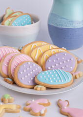 Angled semi-close-up of a plate of yellow, pink and blue decorated homemade cookies with soft-focus bowl and part-jug to the rear, small biscuits on surface to the front.  All on a light background.