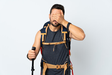 Young mountaineer man with a big backpack and trekking poles isolated on white background covering eyes by hands