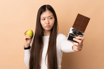 Young Chinese girl over isolated background having doubts while taking a chocolate tablet in one...