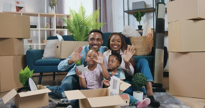 Attractive cheerful happy young african american family with two small kids sitting on the floor in newly acquired apartment among many boxes and waving hands into camera