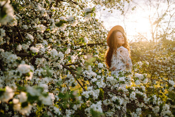 Young woman with beautiful face and long curly hair enjoying scent in blooming spring garden. Spring fashion and lifestyle.