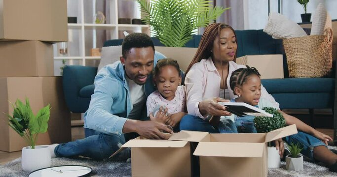 Real estate concept where attractive smiling happy african american family with kids moving into new flat and unpacking carton box wiith home decor