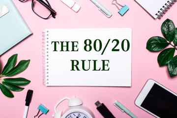 THE 80 20 RULE is written in a white notebook on a pink background surrounded by business accessories and green leaves.
