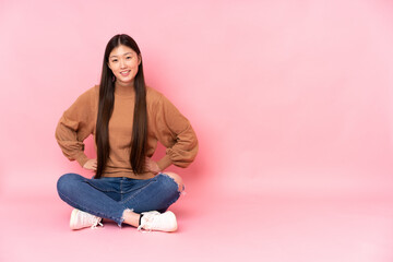 Obraz na płótnie Canvas Young asian woman sitting on the floor isolated on pink background posing with arms at hip and smiling