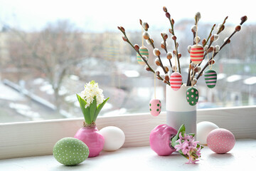Easter decorations on windowsill in Spring. Wooden painted eggs hang on pussy willow twigs in ceramic vase. Easter eggs in pastel colors, pink and green. Urban view over city in window.