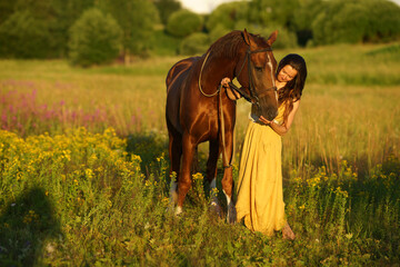 Beautiful woman with long hair in yellow dress standing near brown horse in among purple flowers in green field