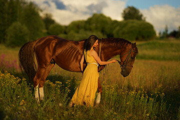Beautiful woman with long hair in yellow dress standing near brown horse in among purple flowers in green field