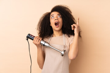 Young african american woman using hand blender isolated on beige background pointing with the index finger a great idea