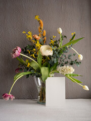 Blank greeting card on a bouquet of spring flowers, white ranunculus, sprigs of yellow forsythia and eucalyptus, pink and white tulips, orange dianthus in a glass vase on a gray concrete background in