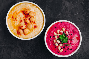 Beet hummus and classic hummus with crisp bread in bowls on dark stone background.
