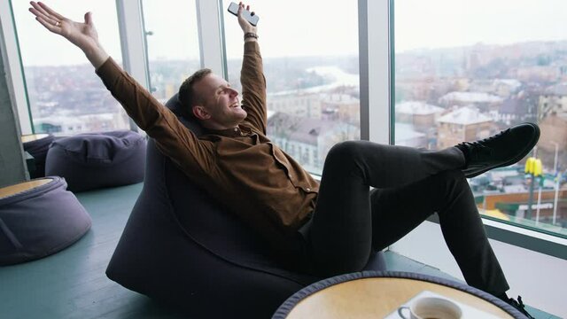 Relaxed businessman indoors. Happy young man is sitting by the window with stretched arms and having a rest in office on the city view window background.
