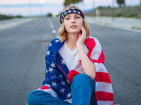 Mujer joven rubia con ojos azules sentada en una carretera celebrando el dia de la independencia de estados unidos