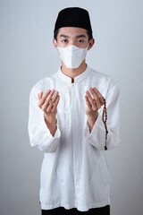 Asian Muslim Man wearing muslim clothes holding prayer beads with medical mask and skull cap praying to God against on grey