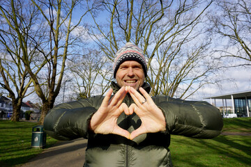Portrait half body of Caucasian handsome man  wearing green jacket, bobble hat with white pom pom making hand to be heart sign with his smiling face in the park 