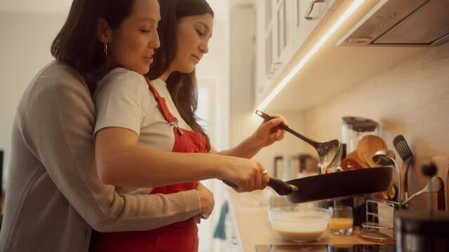 Happy Lesbian Couple Cooking Together in the Kitchen. Girlfriend Embracing Her Partner from the Back while they Prepare Preparing Delicious Meal. Young Partners in Love, Talking, Having Fun. 