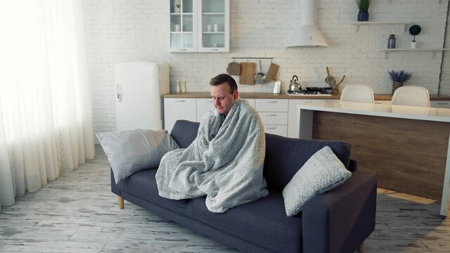 Man having fever. Young man feeling freezy and covering with warm blanket while sitting on a couch at home. Sick man wrapped with blanket during covid outbreak.
