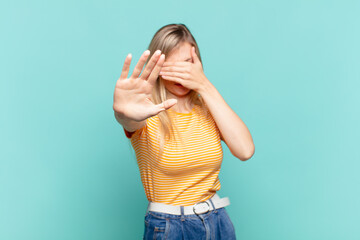 young blond pretty woman covering face with hand and putting other hand up front to stop camera,...