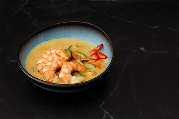 Asian soup with shrimps and vegetables: zucchini, red pepper and carrot. Dish isolated in a blue bowl, close-up on a black marble background. Asian cuisine.