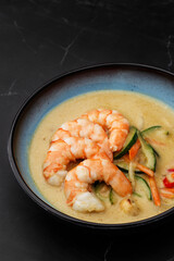 Asian soup with shrimps and vegetables: zucchini, red pepper and carrot. Dish isolated in a blue bowl, close-up on a black marble background. Asian cuisine.