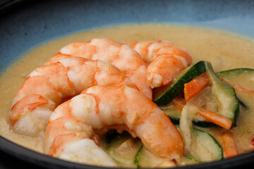 Asian soup with shrimps and vegetables: zucchini, red pepper and carrot. Dish isolated in a blue bowl, close-up on a black marble background. Asian cuisine.