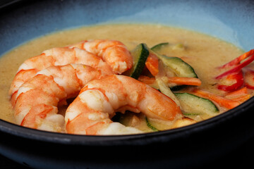 Asian soup with shrimps and vegetables: zucchini, red pepper and carrot. Dish isolated in a blue bowl, close-up on a black marble background. Asian cuisine.
