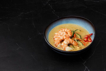 Asian soup with shrimps and vegetables: zucchini, red pepper and carrot. Dish isolated in a blue bowl, close-up on a black marble background. Asian cuisine.