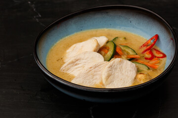 Asian soup with chicken and vegetables: zucchini, red pepper and carrot. Dish isolated in a blue bowl, close-up on a black marble background. Asian cuisine.