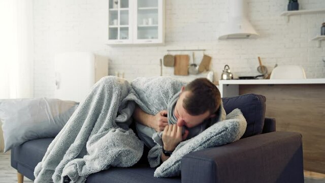Young man trembling from fever. Sick man lying on couch at home and covering with warm blanket over the head.
