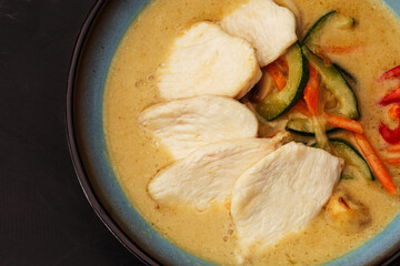 Asian soup with chicken and vegetables: zucchini, red pepper and carrot. Dish isolated in a blue bowl, close-up on a black marble background. Asian cuisine.