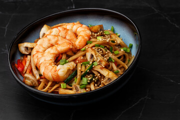 Udon noodles with shrimps and vegetables: zucchini, red pepper, mushrooms, carrot, spring onion and sesame seeds. Dish isolated in a blue bowl, close-up on a black marble background. Asian cuisine.