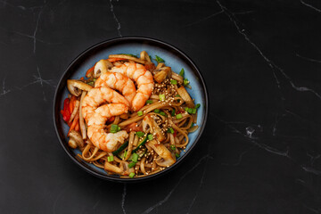 Udon noodles with shrimps and vegetables: zucchini, red pepper, mushrooms, carrot, spring onion and sesame seeds. Dish isolated in a blue bowl, close-up on a black marble background. Asian cuisine.