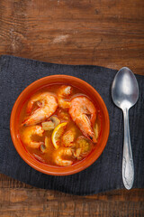 Tom yam soup with shrimps in a plate on a black stand with a spoon on a wooden table.