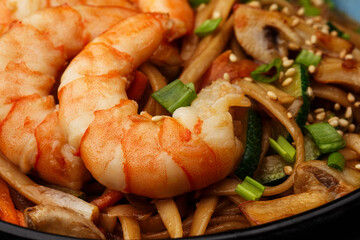 Udon noodles with shrimps and vegetables: zucchini, red pepper, mushrooms, carrot, spring onion and sesame seeds. Dish isolated in a blue bowl, close-up on a black marble background. Asian cuisine.