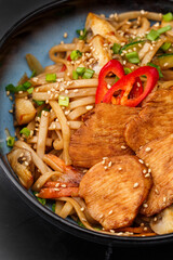 Udon noodles with teriyaki chicken and vegetables: zucchini, red pepper, mushrooms, carrot, onion and sesame seeds. Dish isolated in a blue bowl, close-up on a black marble background. Asian cuisine.